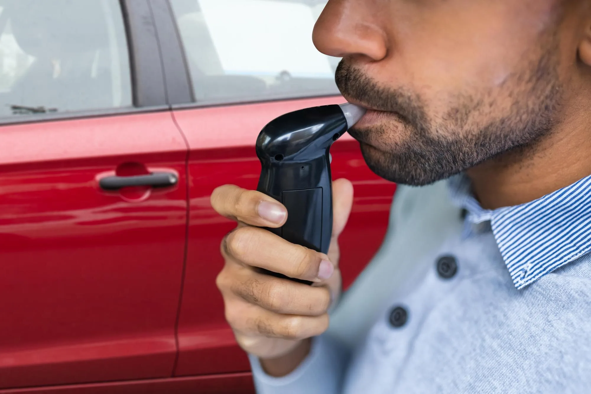 Teste de bafômetro. Na imagem temos um carro vermelho ao fundo e a frente, um homem assoprando o bafômetro. Este homem veste uma camisa de botões na cor azul clara, tem a pele morena e pouca barba.